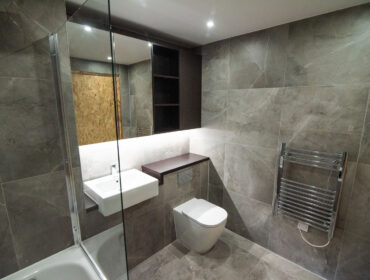 Grey bathroom with a bathtub behind a glass screen, a wall-hung toilet and a gold-accented niche.